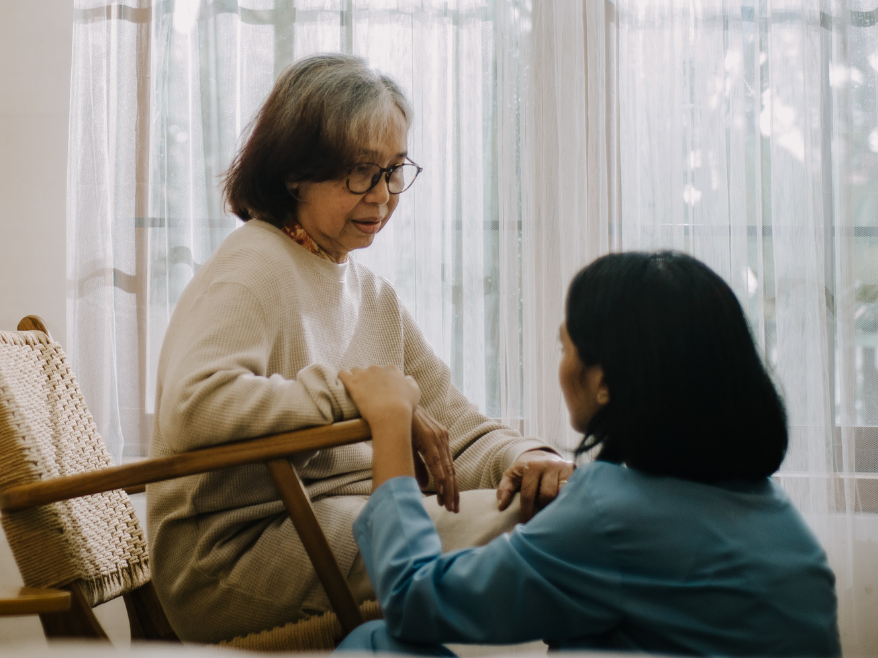 A senior-aged woman with hair that is blaack with some white mixed in, sits in a chair while another woman kneels on the ground in front of her holding her hand.